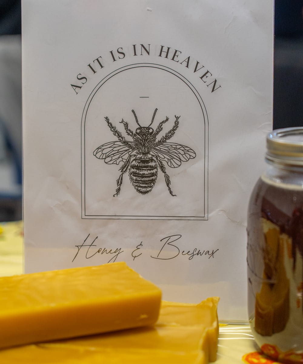 The As It Is In Heaven sign on Paxton's market table, an engraved honeybee inside an arch, with slabs of beeswax and a jar of honey in front of it
