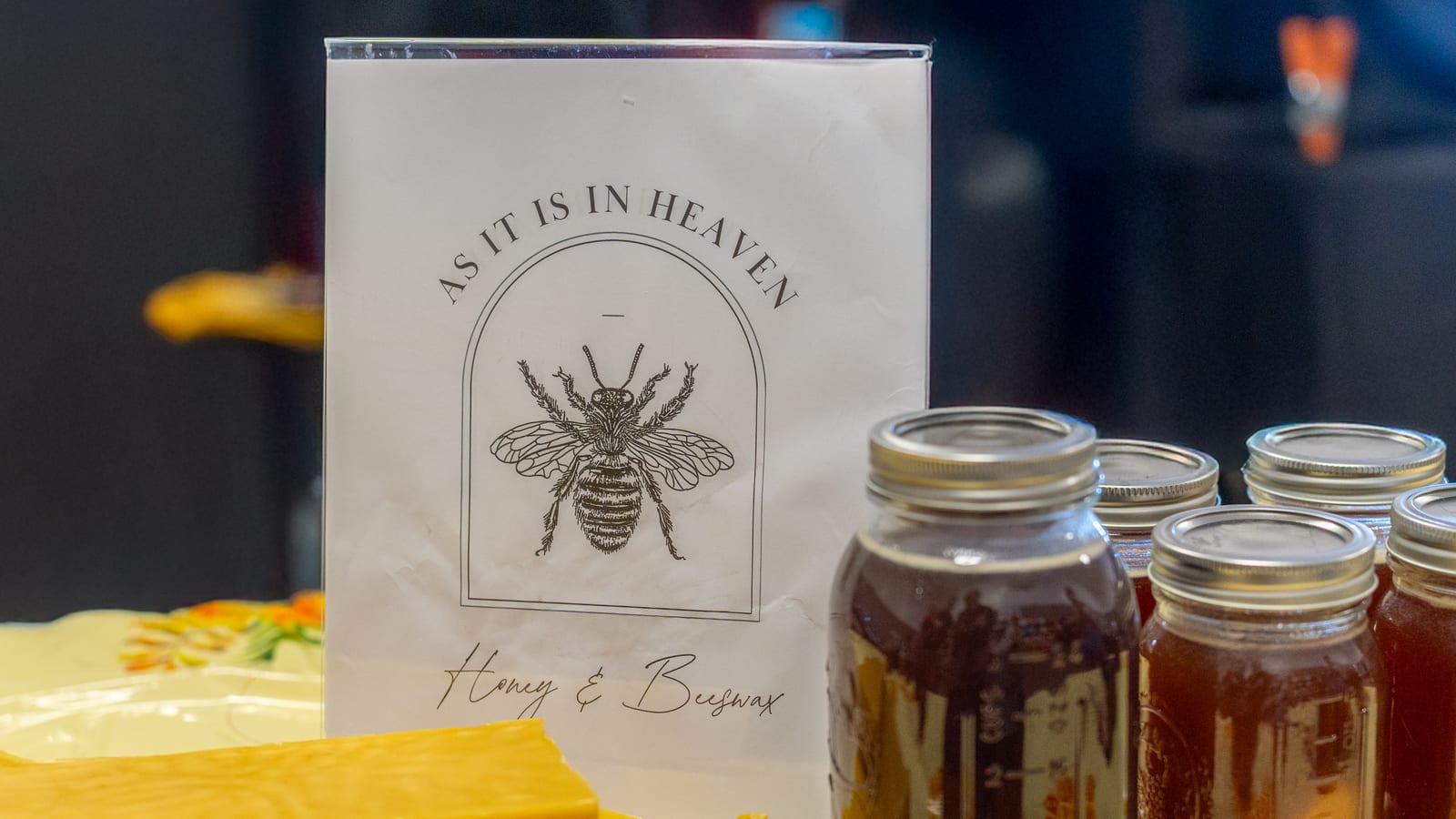 The As It Is In Heaven market sign, an engraved honeybee inside an arch above the words Honey and Beeswax, standing behind jars of honey