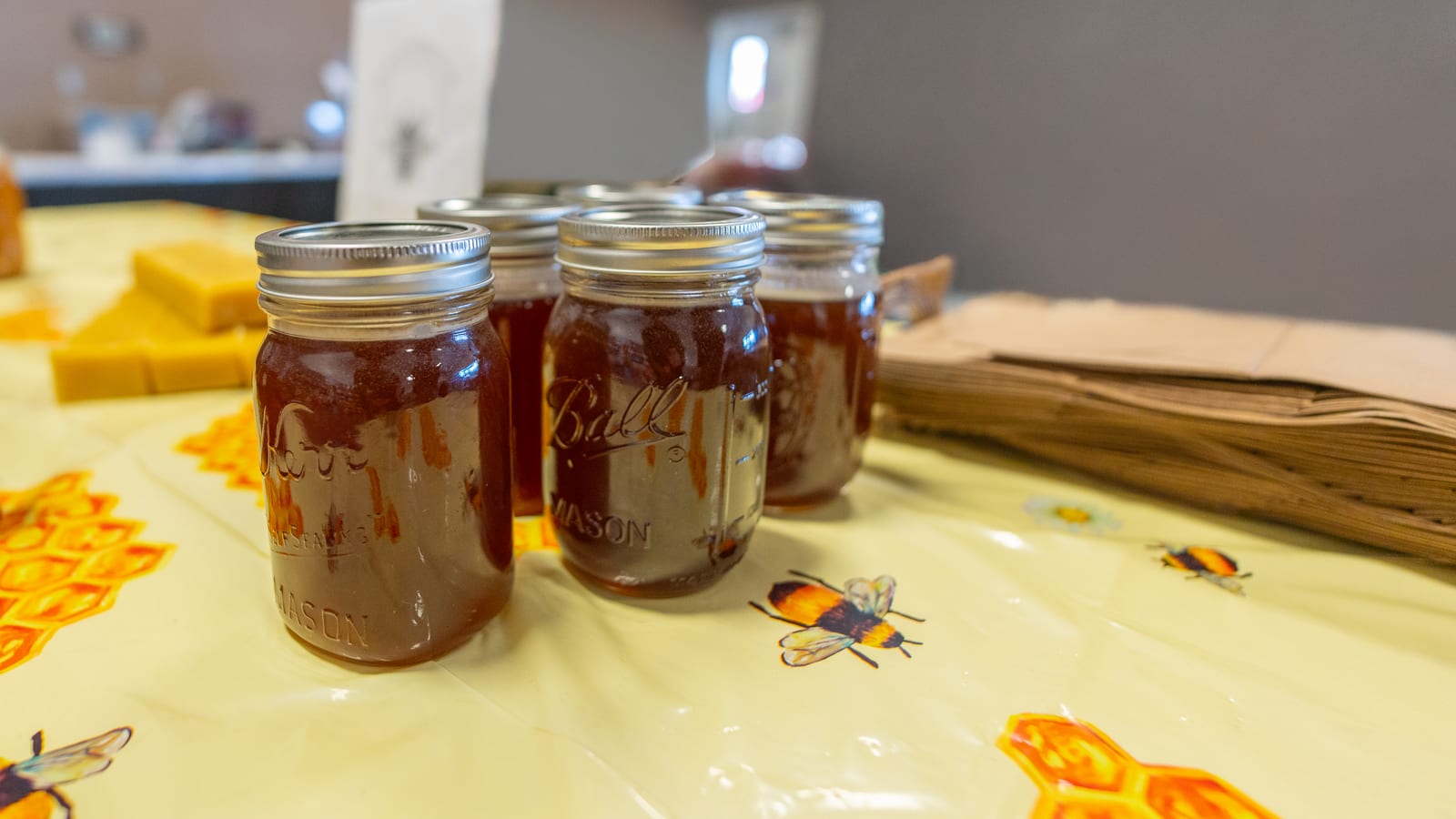 Sealed mason jars of dark honey on a bee-print market table, with blocks of beeswax and a stack of paper bags beside them