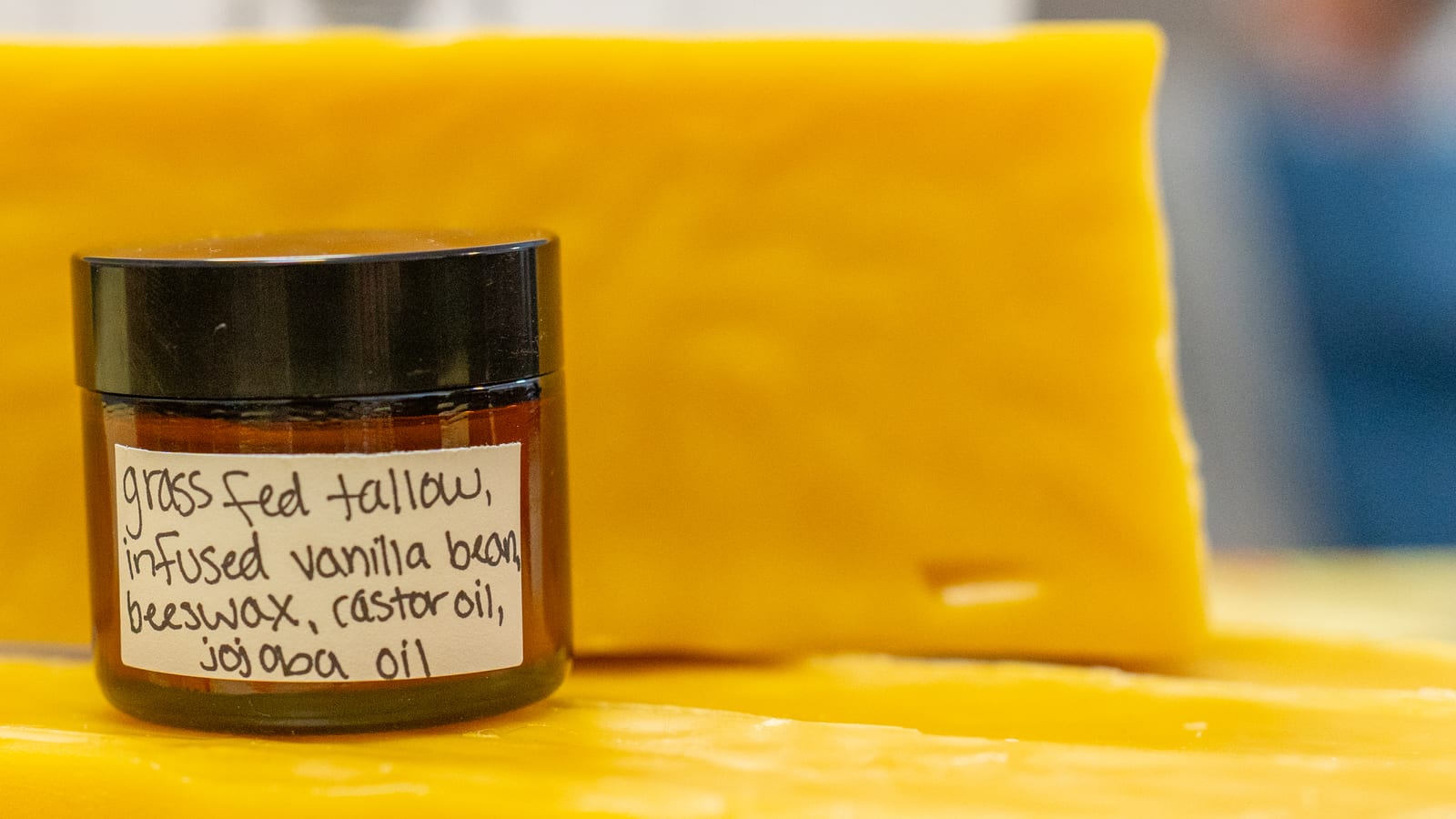 A close view of an amber jar of tallow lotion on a slab of beeswax, with a handwritten label listing grass fed tallow, infused vanilla bean, beeswax, castor oil and jojoba oil