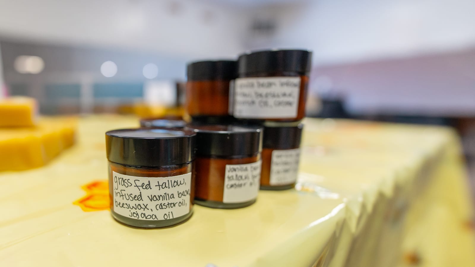 Five small amber jars of tallow lotion on a bee-print market cloth, the nearest one labeled grass fed tallow, infused vanilla bean, beeswax, castor oil, jojoba oil