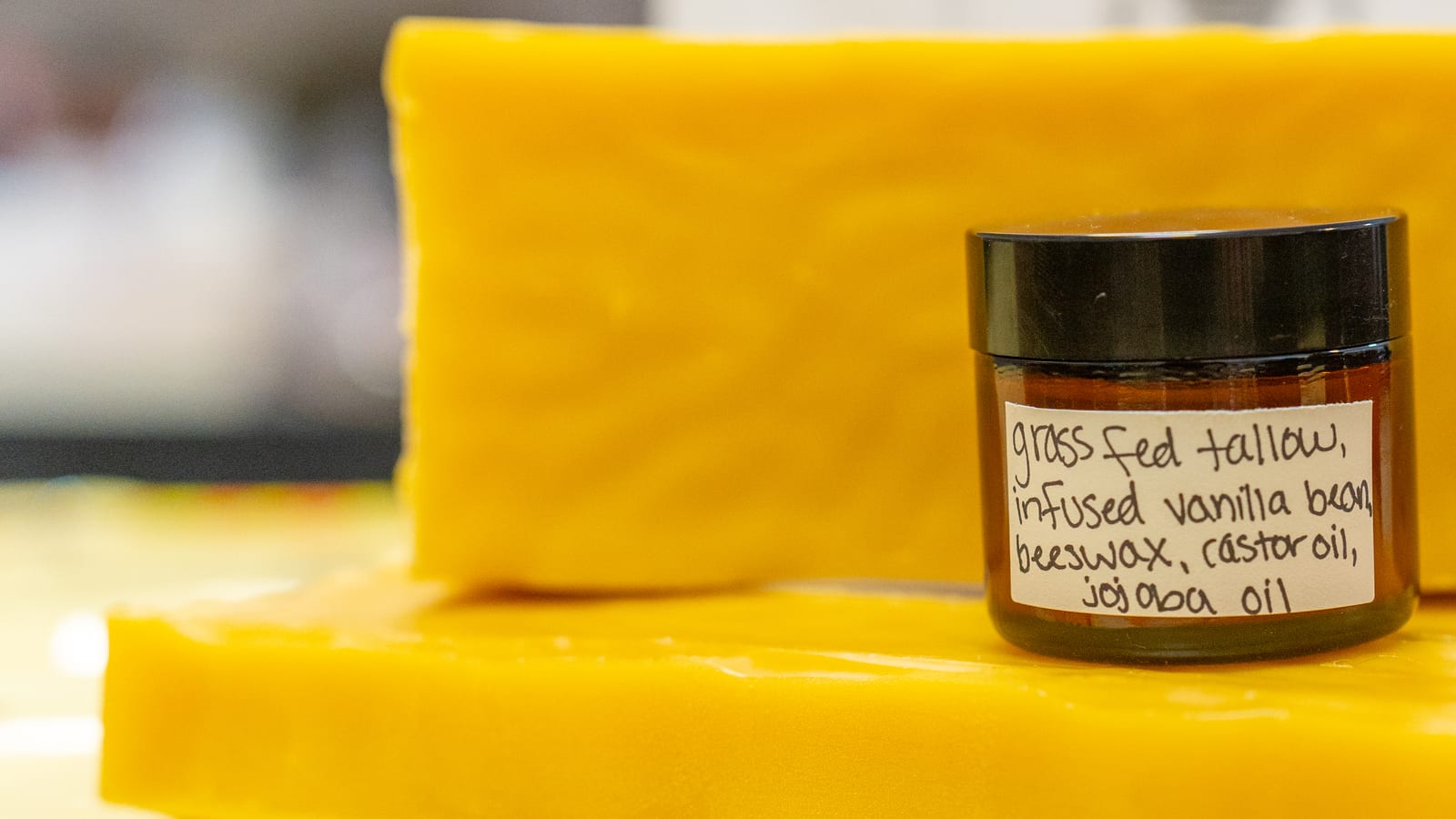 A jar of whipped tallow lotion labeled grass fed tallow, infused vanilla bean, beeswax, castor oil, jojoba oil, standing on slabs of beeswax