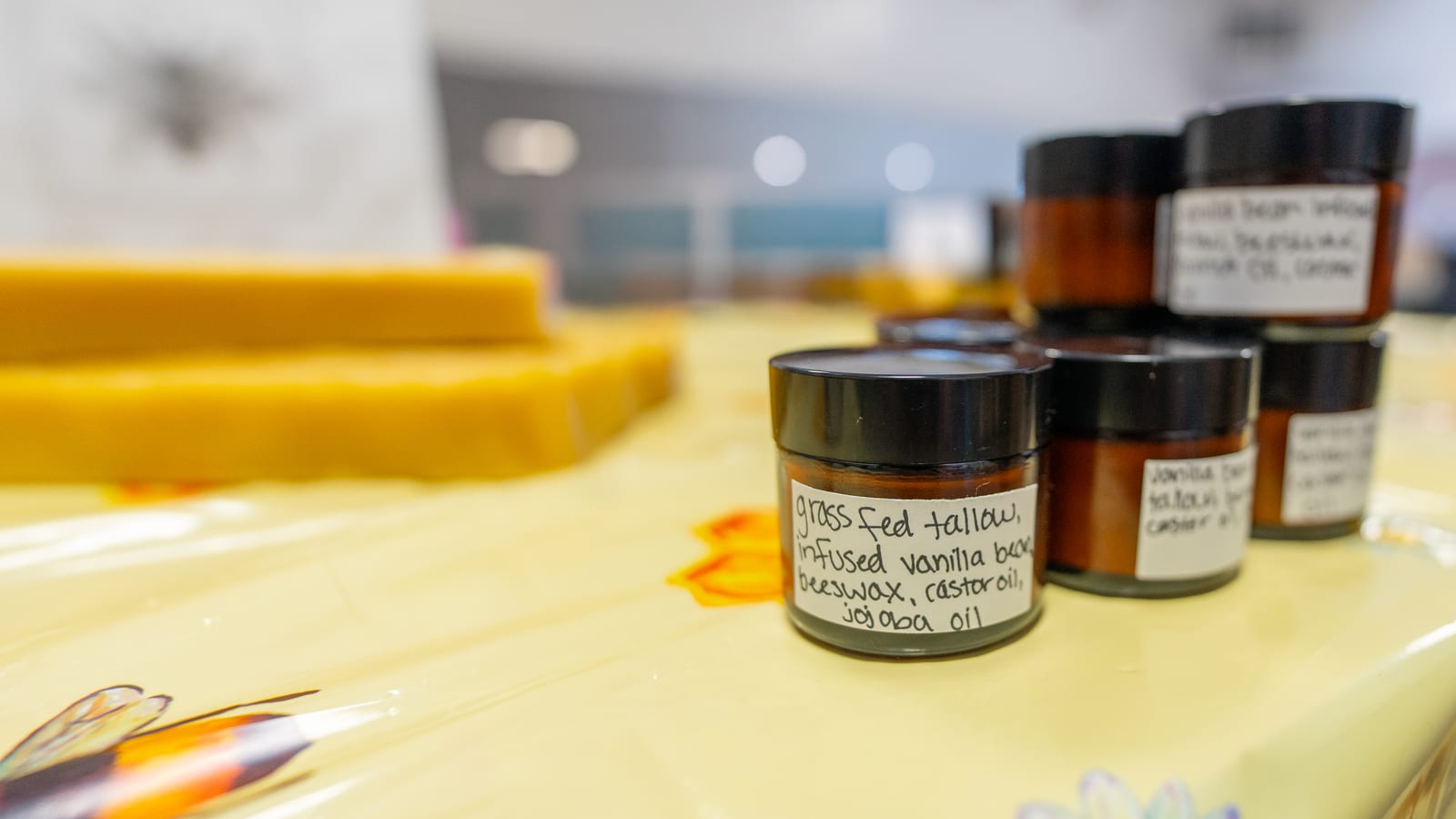 Slabs of golden beeswax next to a row of small amber jars of tallow lotion on a bee-print market cloth