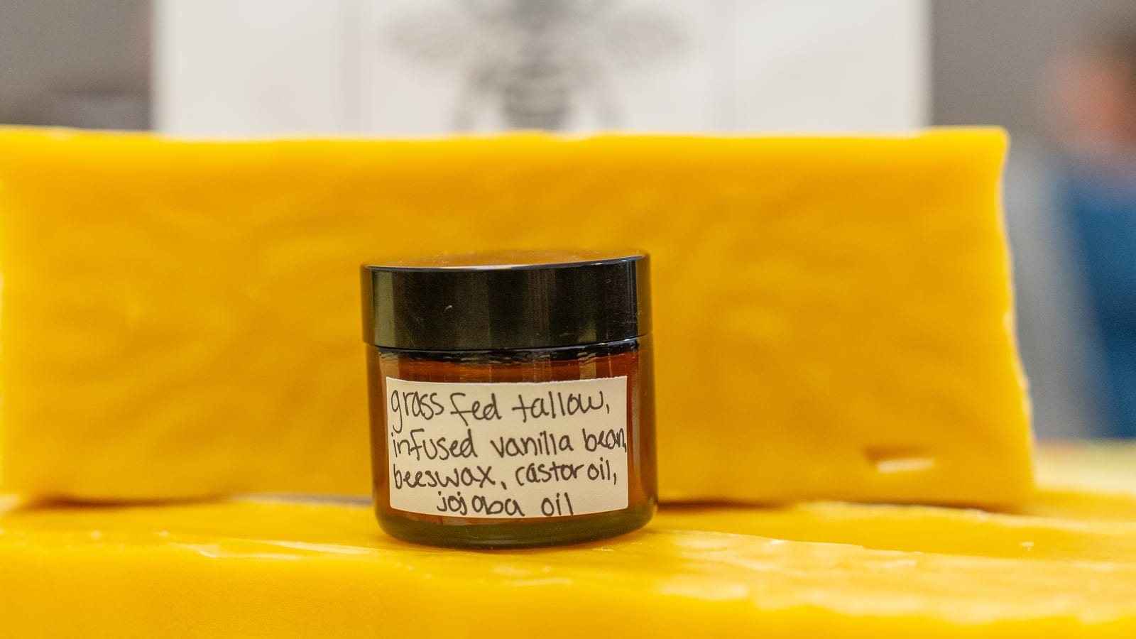 A small amber jar of tallow lotion resting on a slab of beeswax, its handwritten label reading grass fed tallow, infused vanilla bean, beeswax, castor oil, jojoba oil