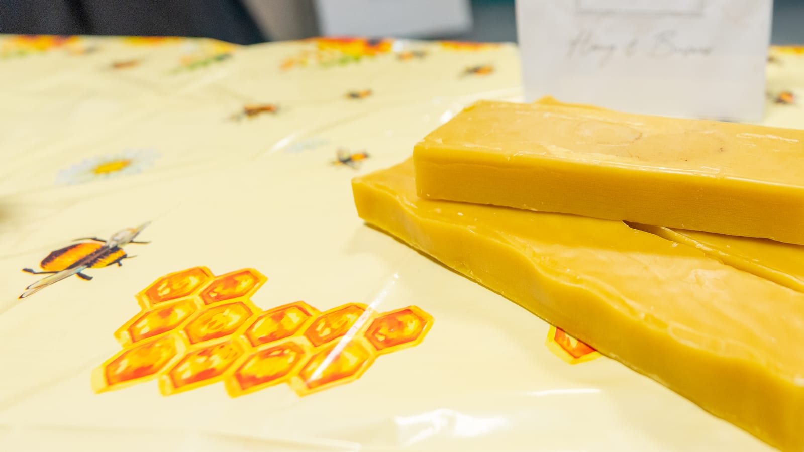 Two poured slabs of golden beeswax on a bee-print market cloth, with the As It Is In Heaven sign behind them