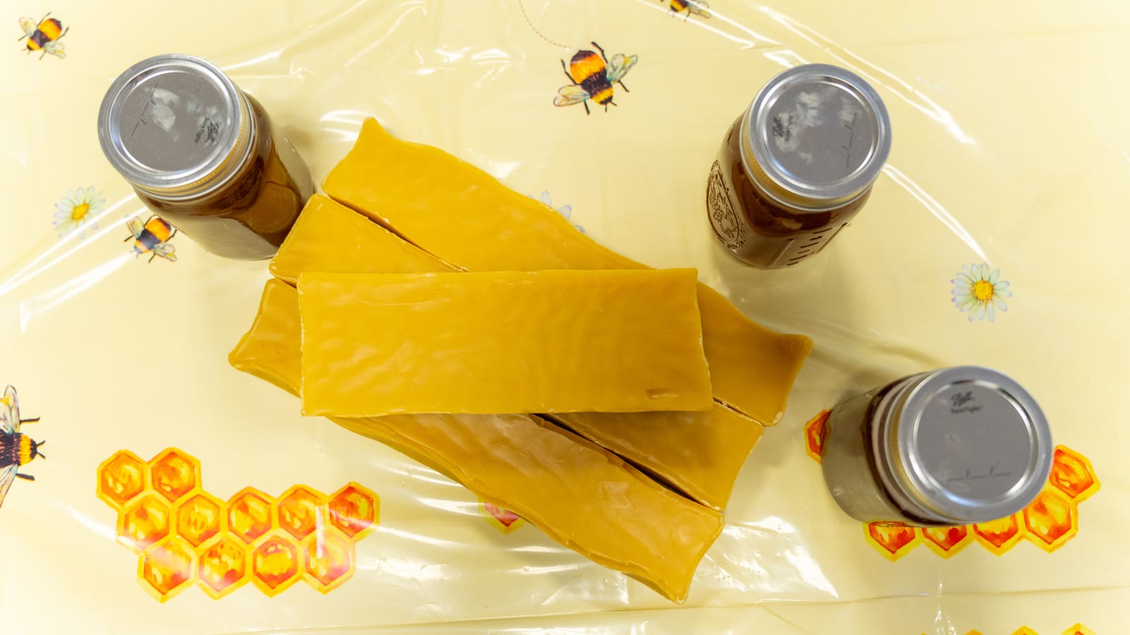 Three bars of golden beeswax laid out on a bee-print cloth with the lids of three honey jars beside them