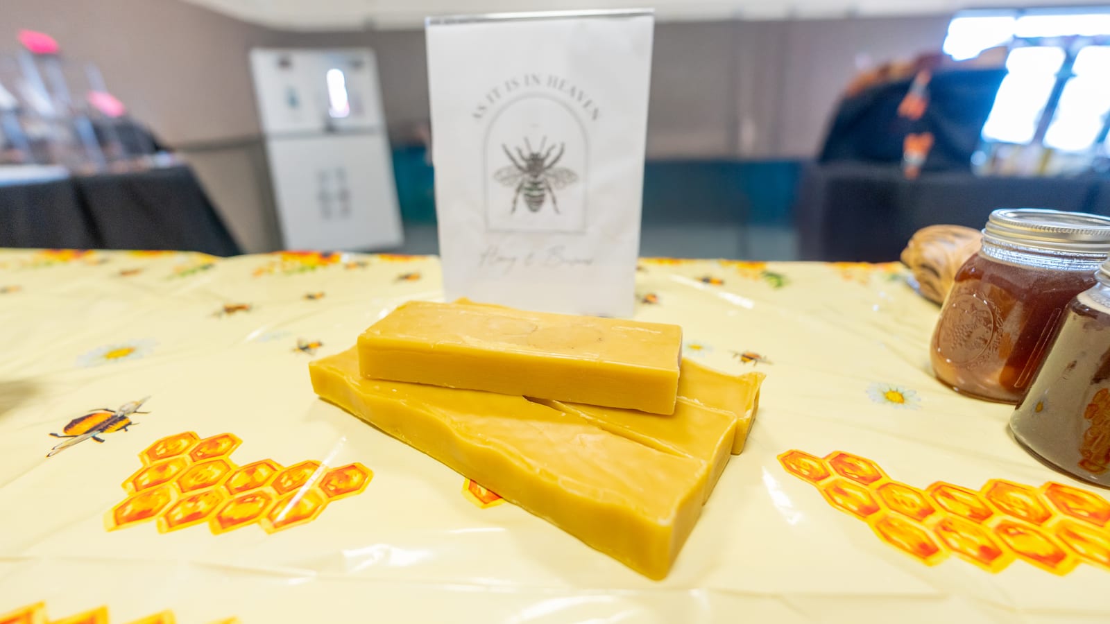 Two poured slabs of golden beeswax on a bee-print market cloth, with the As It Is In Heaven sign and a jar of honey behind them