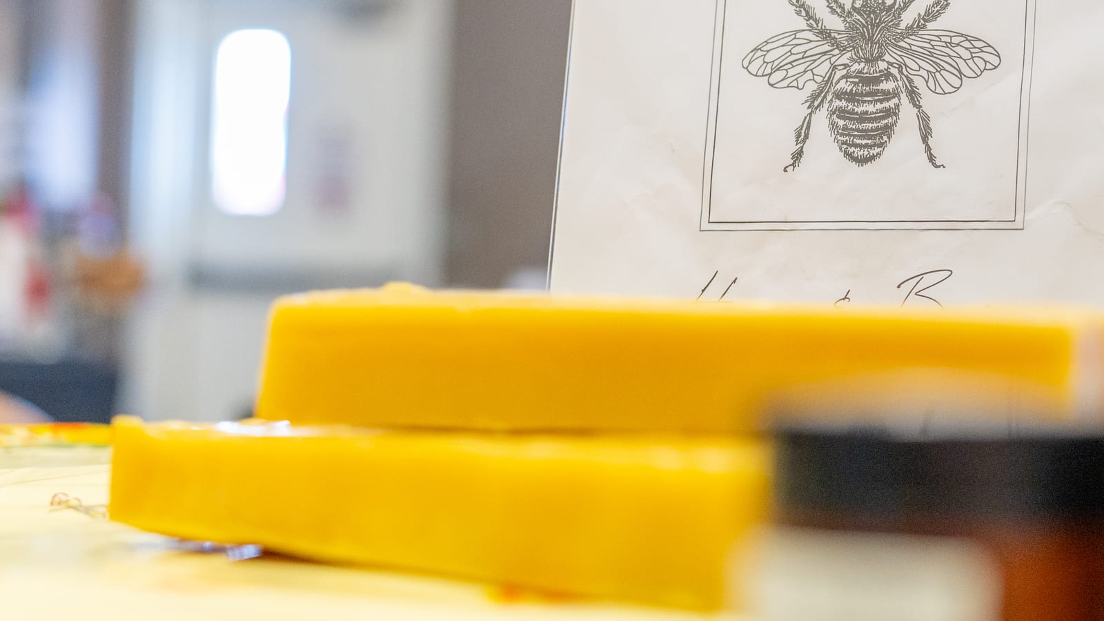 Two poured slabs of golden beeswax stacked on a market table with the As It Is In Heaven sign behind them